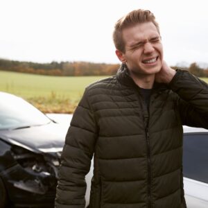 a man holding his neck in pain with a car accident behind him