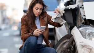 persona que usa su teléfono mientras está sentada cerca de su automóvil dañado después de un accidente automovilístico