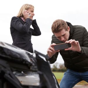 Man photographing a car accident while a woman stands next to him on the phone. 