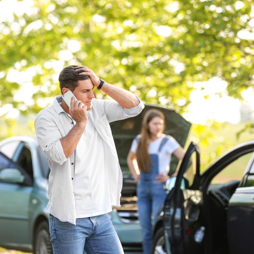 Man looking distressed on the phone at the scene of a car accident with a woman standing in the background. 