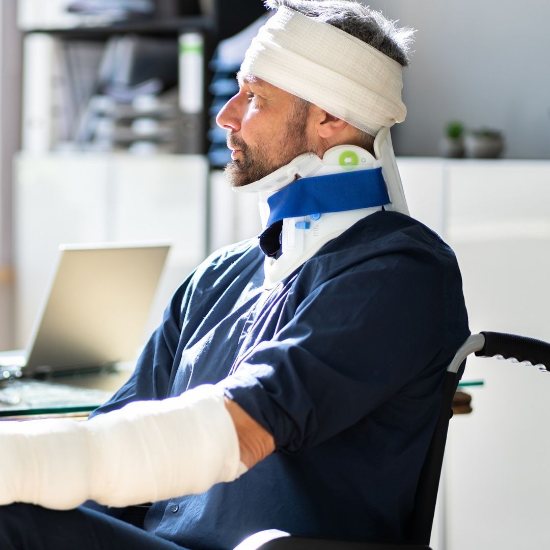 Injured man sitting in a wheelchair with a head bandage, neck collar, and arm cast.